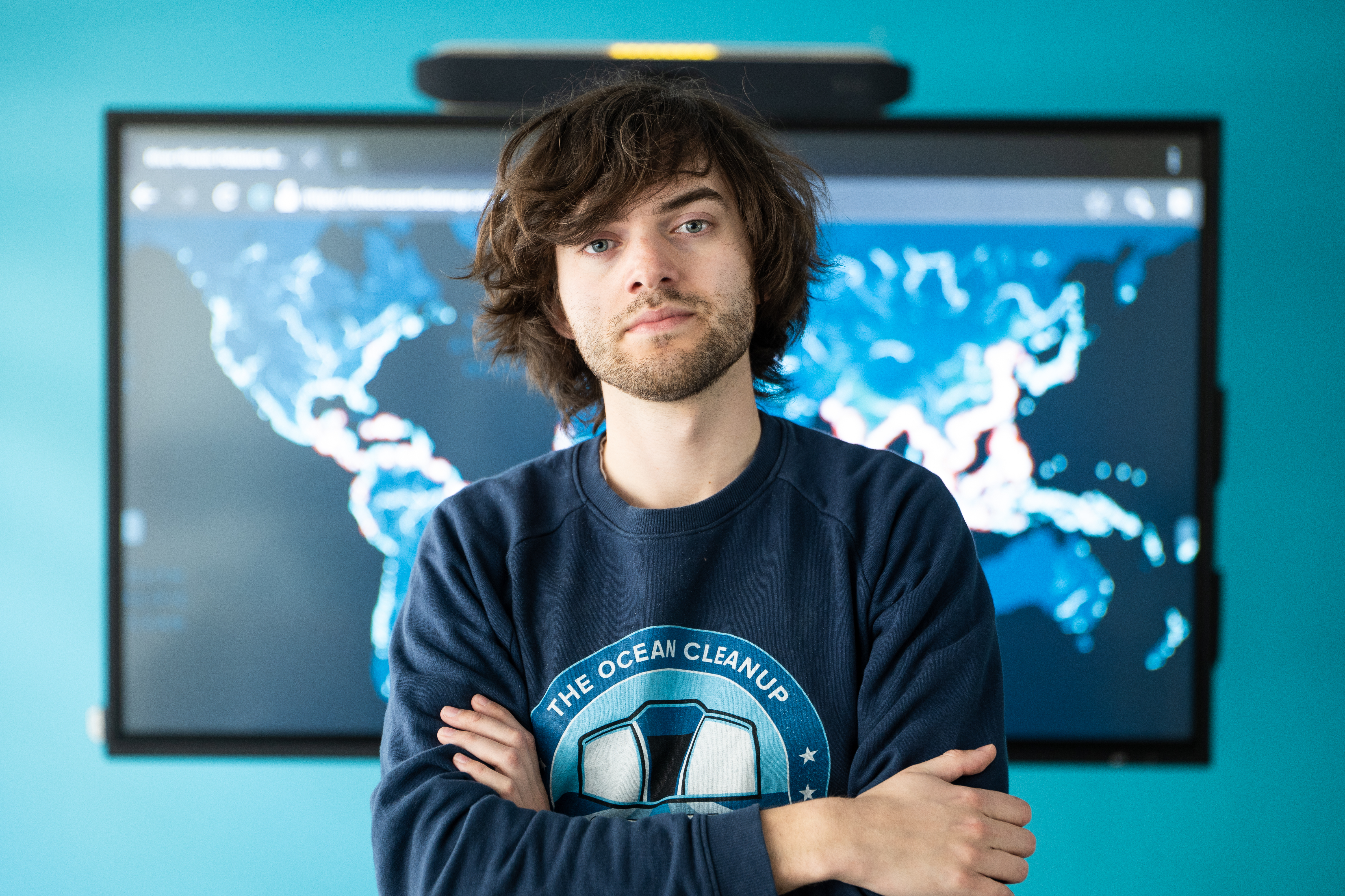 Bayan Slat de The Ocean Cleanup Project, vestido con una sudadera azul marino, se encuentra frente a un monitor que muestra un mapa mundial.