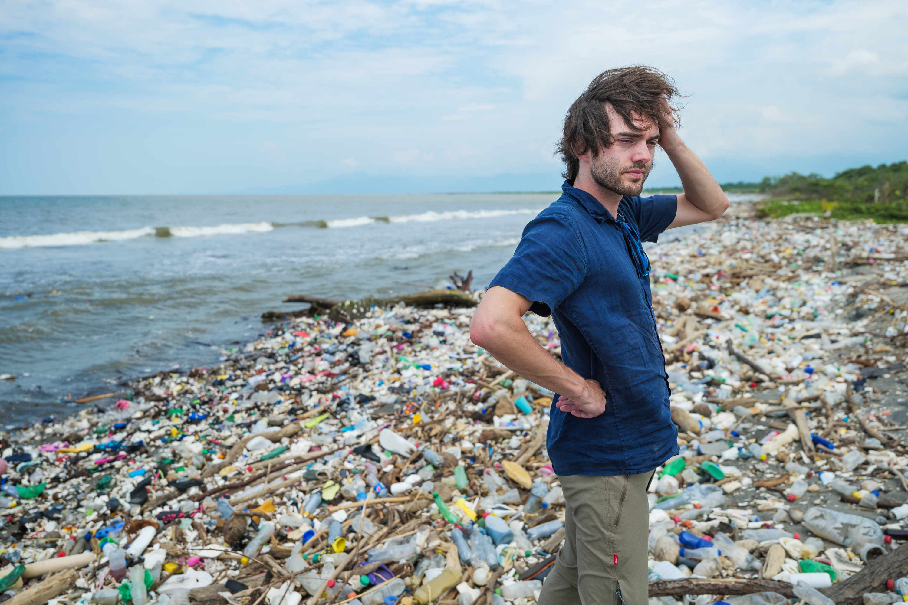 Un hombre que parece molesto parado en una playa llena de botellas de plástico y escombros.