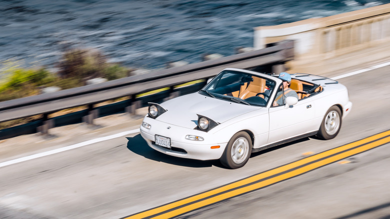 Mazda Miata blanco conduciendo por la carretera costera, ángulo alto, vista frontal 3/4, interior color canela