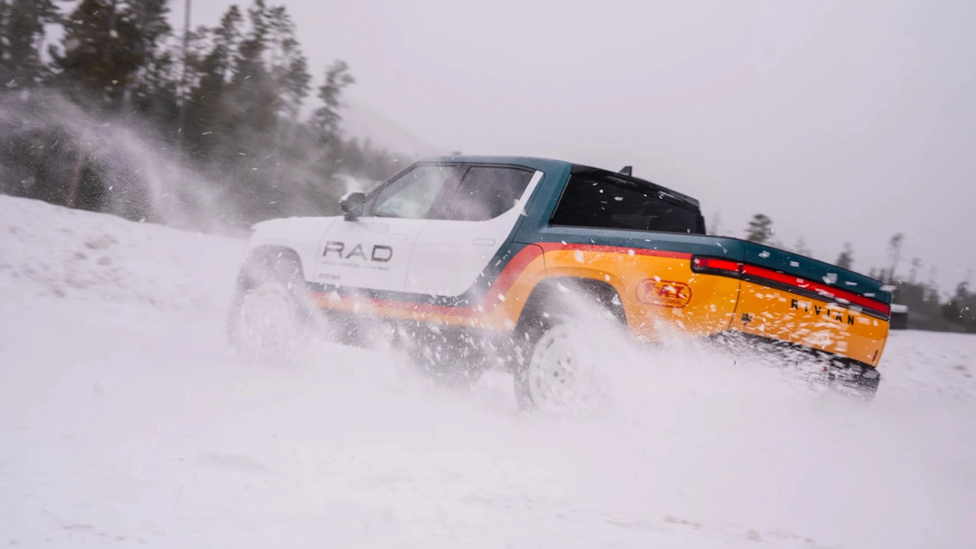 Camioneta Rivian multicolor conduciendo a través de la nieve con nieve rociada alrededor de las ruedas.