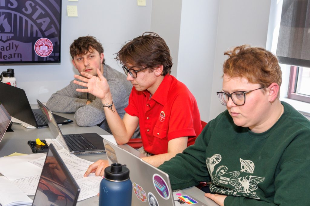 Tres estudiantes conversan en una mesa con computadoras portátiles en un aula de la Universidad Estatal de Illinois.