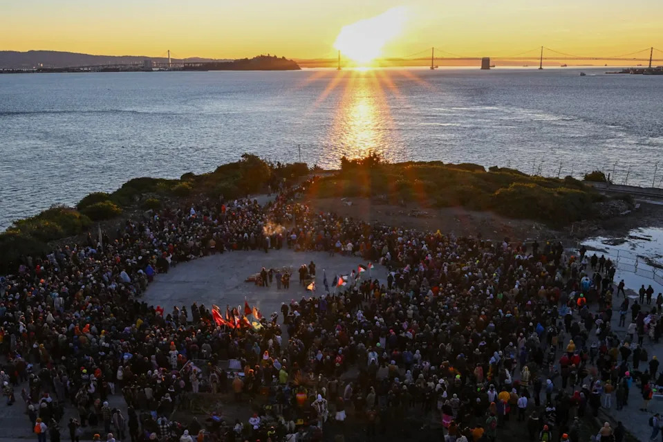 Un grupo de personas se encuentra alrededor de un círculo abierto en una isla, mientras el sol sale detrás de un puente sobre el agua.