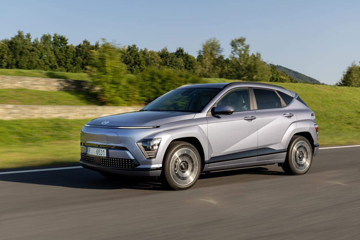 Un Hyundai Kona Electric plateado circula por la carretera. Pasa por un campo de hierba y algunos árboles que dan sombra.