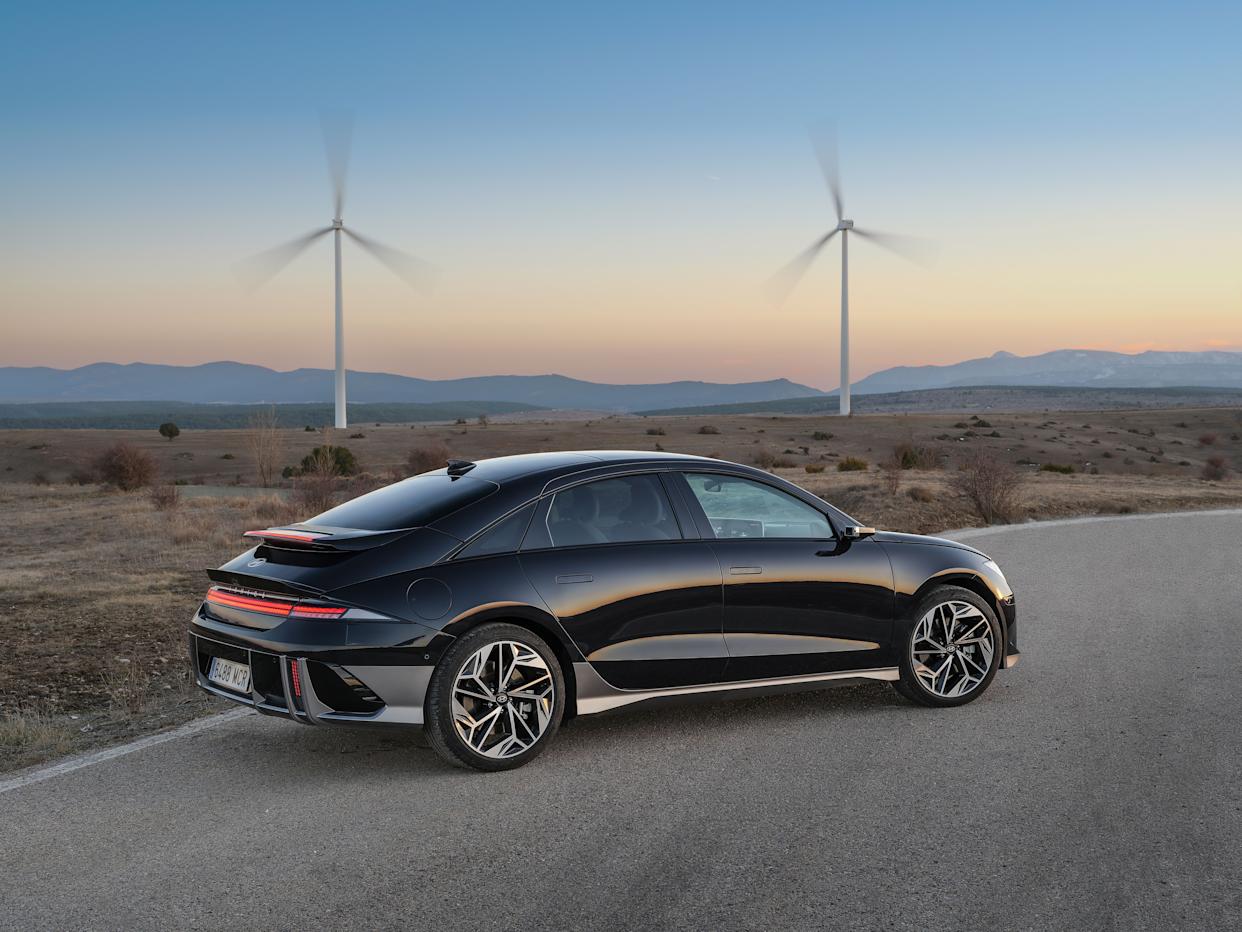 Un sedán Hyundai Ioniq 6 negro está estacionado en una extensión de alquitrán junto a un gran campo de arena. El sol se pone detrás de montañas lejanas y hay dos molinos de viento girando.