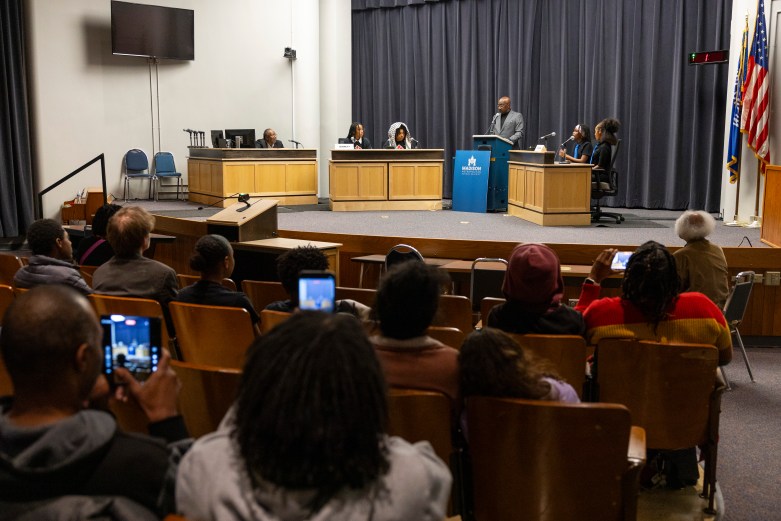 Una audiencia se sienta frente a un escenario donde varias personas se sientan detrás de escritorios con micrófonos mientras otra persona se encuentra en un podio etiquetado 