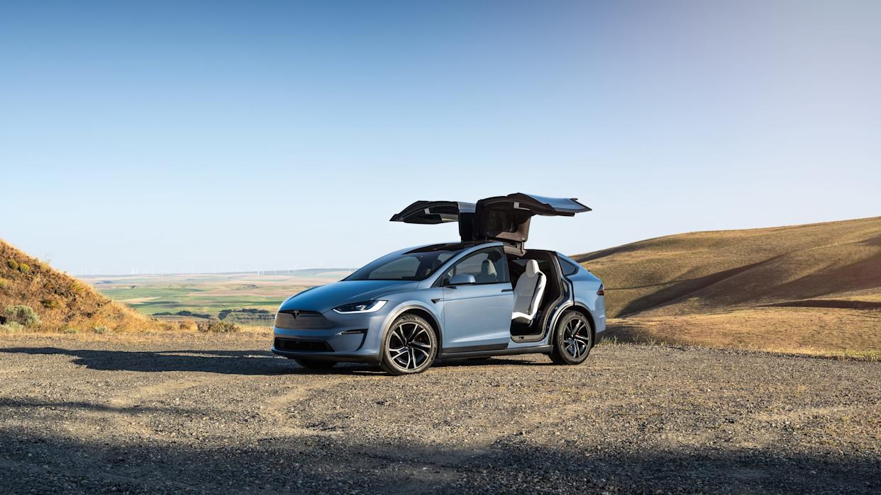 Un Tesla Model X azul claro está estacionado en el lado de grava de una colina arenosa. Se abren las puertas traseras en forma de alas de gaviota.